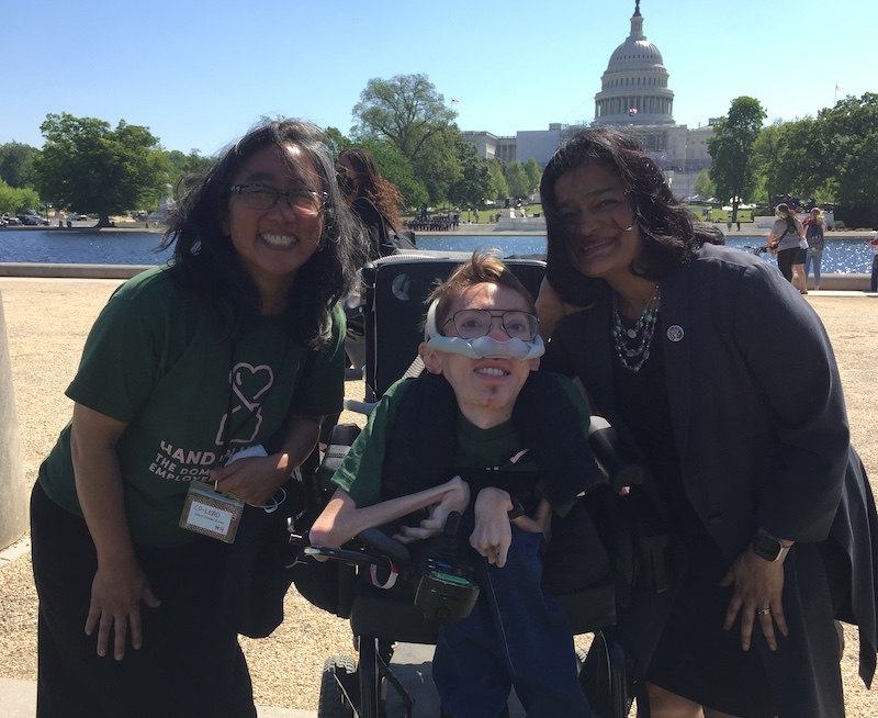 White man in wheelchair and a breathing device smiles in front of Washington Capital with two women of color on either side.
