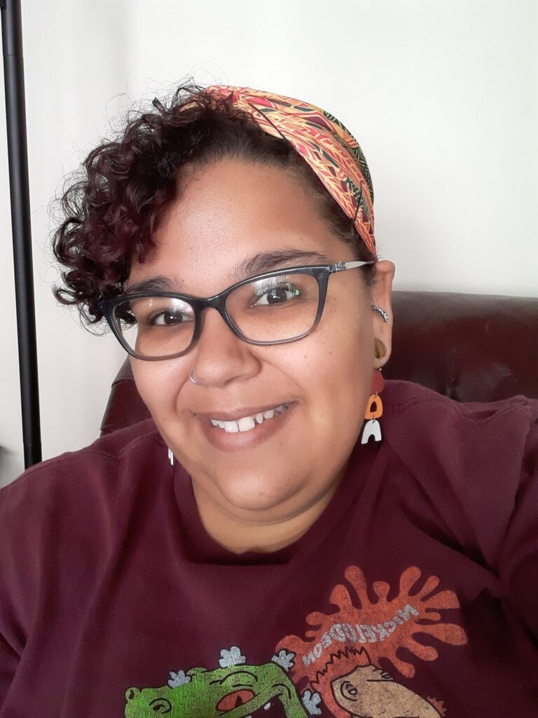 A medium-toned Black woman with glasses and short curly brown hair wearing a Kenta cloth hair wrap, dangle earrings, and a red t-shirt is smiling towards the camera