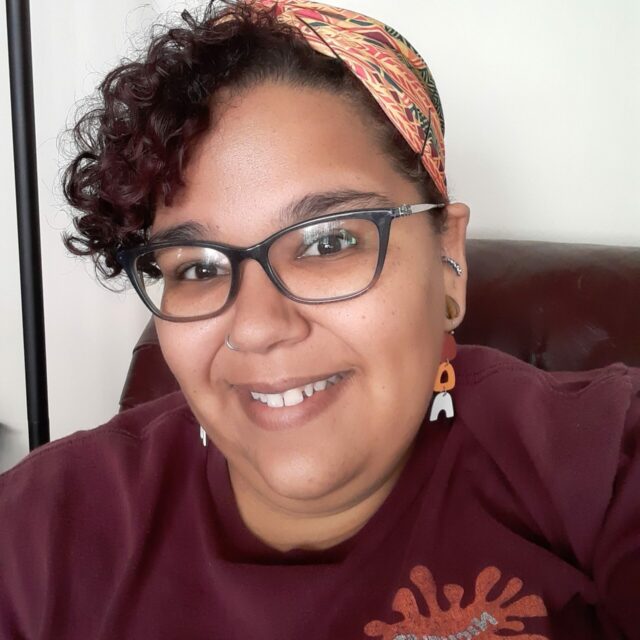 A medium-toned Black woman with glasses and short curly brown hair wearing a Kenta cloth hair wrap, dangle earrings, and a red t-shirt is smiling towards the camera
