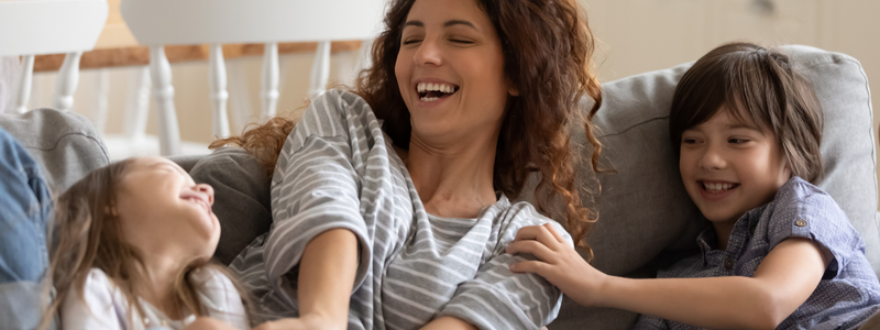 A white woman on the couch laughing with curly brown hair with two other small children laughing as well. A little white girl on the left laughing, and a little white boy with short brown hair is tickling the woman. 