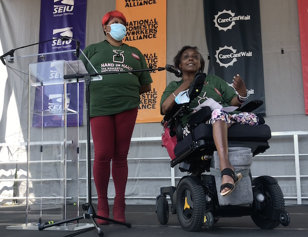 black women with burgundy pants and boots, and green Hand in Hand shirt standing on stage with hands behind back, next to a women sitting win wheelchair with short curly black hair and green honor domestic work shirt speaking. 