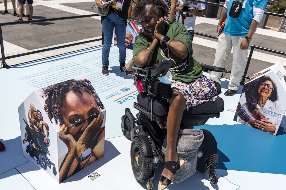 photo of Engracia looking at square sculpture of herself, while sitting in wheelchair, with green hand in hand shirt. While she holds her head in the palm of her hands with a shocked expression.