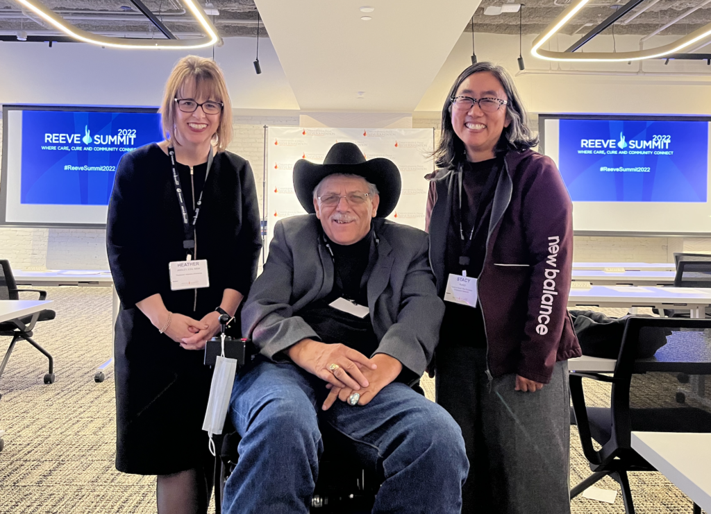 3 people in a room white woman on the left with short blonde hair and bangs smilling while hands are folded in the front of her. White man with black hat and grey suit coat and blue denim jeans sit in wheelchair. Asian women with short black hair and glasses and smiling towards the camera.