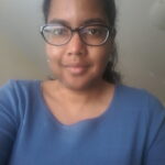 A black woman with black hair pulled into a ponytail while wearing black glasses and a blue shirt.