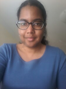 A black woman with black hair pulled into a ponytail while wearing black glasses and a blue shirt. 