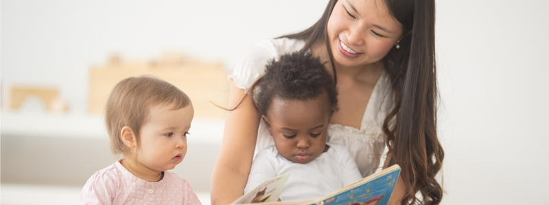 An asian woman is holding a blue book outside, while a black baby is sitting on her lap and another white baby is sitting close, looking at the book.
