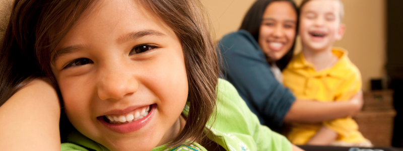 A young white girl smiling into the camera while a black woman and a young white boy are smiling and hugging in the background.