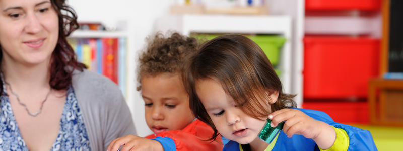 A white woman with dark brown hair watching two babies playing. The baby on the right is white with dark brown hair and a blue shirt. The baby on the left has light brown skin and curly brown hair wearing a red shirt. 