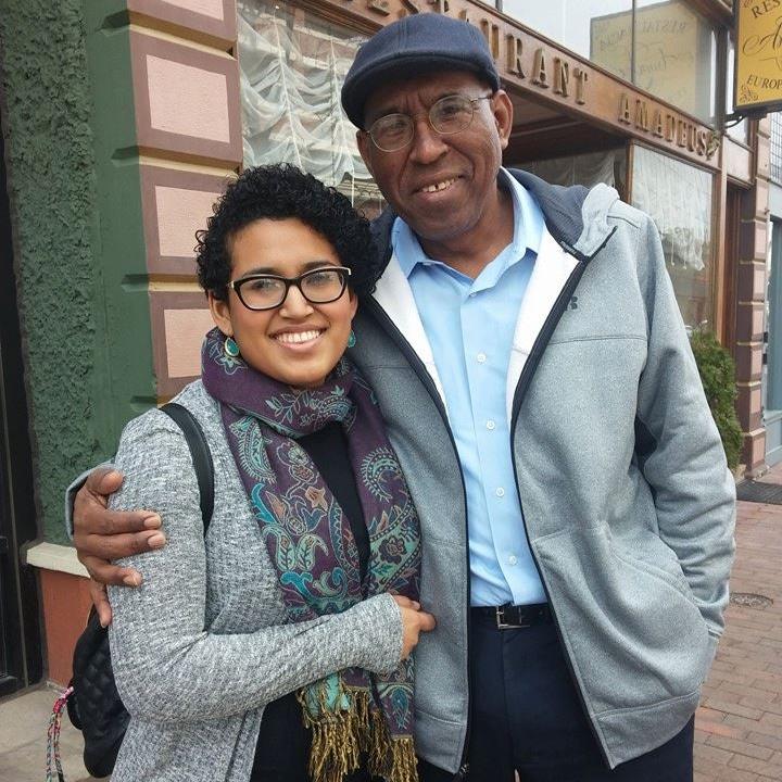 Afro-Latinx Man wearing glasses, a dark blue flat cap hat and a gray hoodie over a light blue dress shirt, left hand in his pant's pocket, his right arm around his Afro-latinx offspring, Veralucia, wearing glasses, a scarf with purple and green designs around their neck, and a gray cardigan, black bag drapped over shoulder. Both smiling widely at the camera. Business fronts in the background.