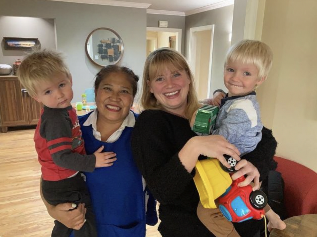 Two women, one, a woman of color with hair pulled back, wearing a blue top with white collar, smiling widely, as she holds a young white child with blonde hair, wearing a gray and red long-sleeve shirt looking at the camera. She is standing next to a white woman with blonde hair, wearing a black top, holding another young white child with blonde hair, wearing a blue and white shirt, and holding a red and yellow toy truck. Furniture and a wall mirror in the background.]