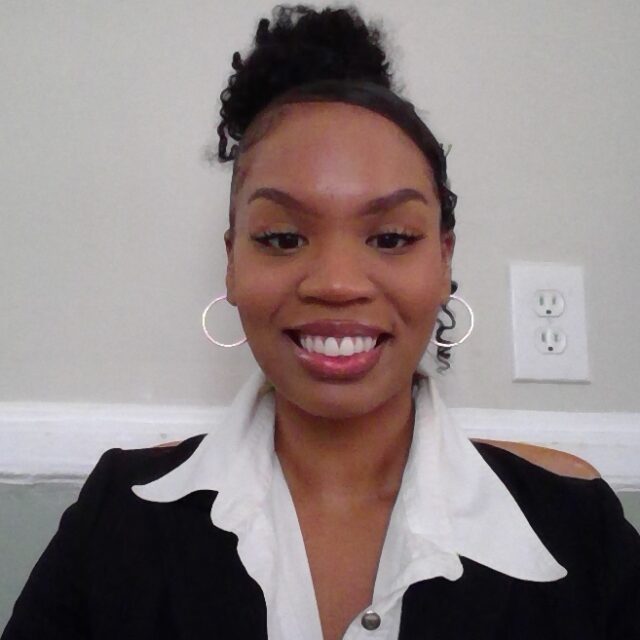 A headshot of a black women wearing a black shirt with a wide white collar, and black curly hair tied into a bun, while smiling into the camera in front of a wall.