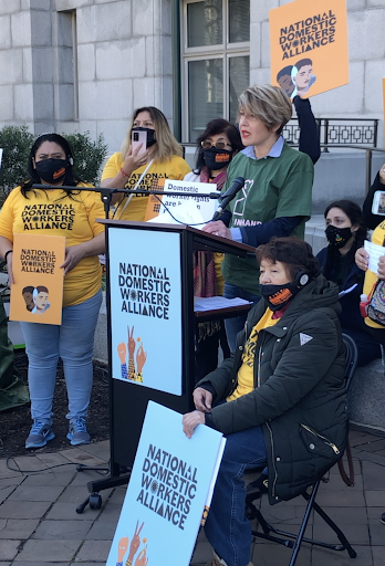 A white woman with blonde hair standing at podium outside speaks with a green Hand in Hand shirt. There are women to her left holding National Domestic Workers Alliance posters, and a woman on the right sitting in a chair holding a poster. 