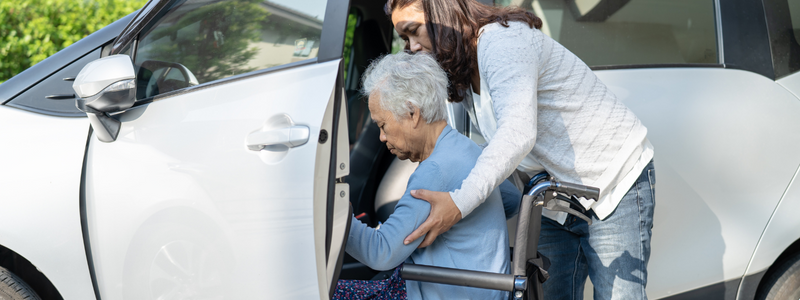 Photograph of an older woman seated in a wheelchair transferring into a white vehicle with assistance from a woman from behind. 