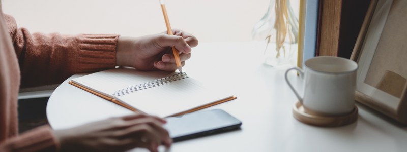 Photograph of an open spiral-bound journal being written in by a black woman holding a pencil. Beside the journal sits a phone, also being used by the woman. On the right side of the image sits a white coffee cup on top of a wooden coaster. 