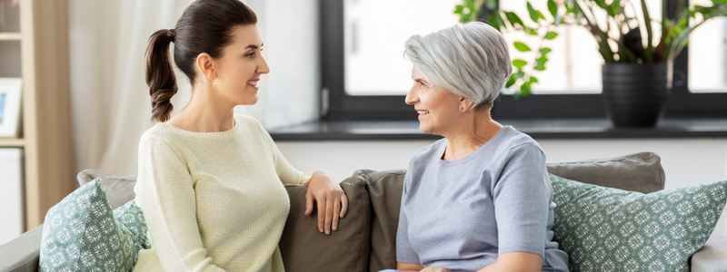 Photograph of two women conversing while sitting on a sofa. On the left is a woman with dark hair wearing a pale yellow sweater. On the right is an older woman with gray hair wearing a light blue-gray shirt. 