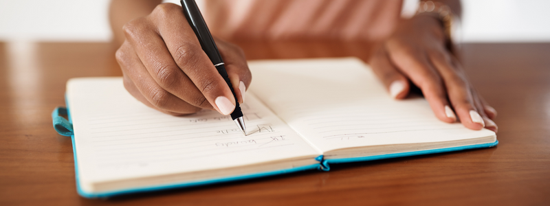 A black woman holding a pen writes a series of tasks in a journal.