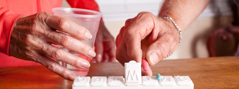 Two people shown in hand only preparing to take medication. On the left a light skinned hand holds a cup of water and on the left a light skinned hand of another person holds a pill. 