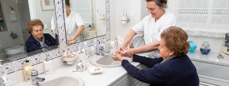 A light skinned older woman is being assisted washing her hands in a bathroom sink by a light skinned caregiver.