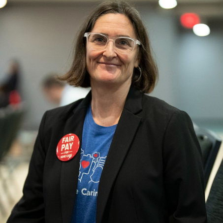 Headshot of a white woman wearing clear glasses with brown hair sitting in an hall wearing a black blazer with blue shirt, and red pin on the left of the blazer.