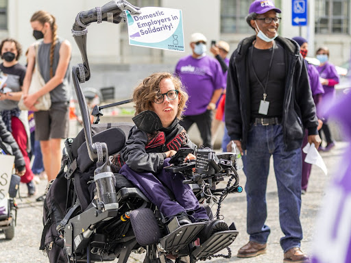 Person in a wheelchair with an mechanical arm that holds a sign that says domestic employers in solidarity!