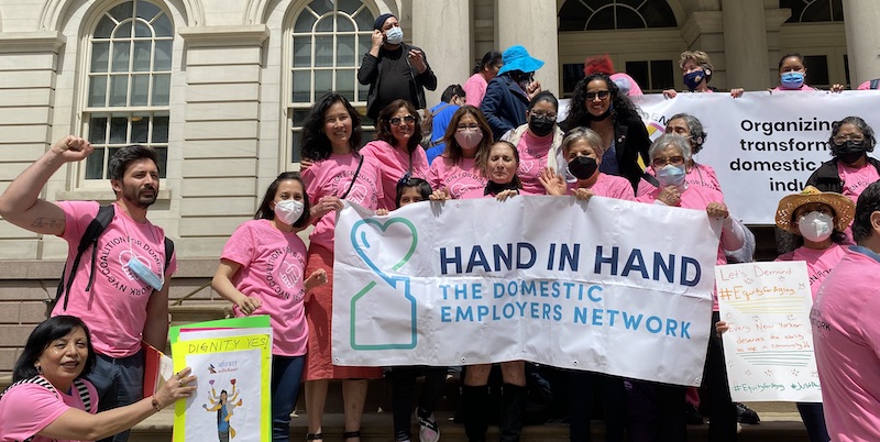 Group of people outside city hall steps wearing pink shirts that say NYC domestic worker coalition and holding a Hand in Hand Banner