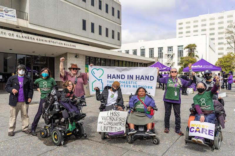 Group photo of people both sitting in wheelchairs and standing outside while wearing green Hand in Hand shirts and holding posters in support of IHSS.
