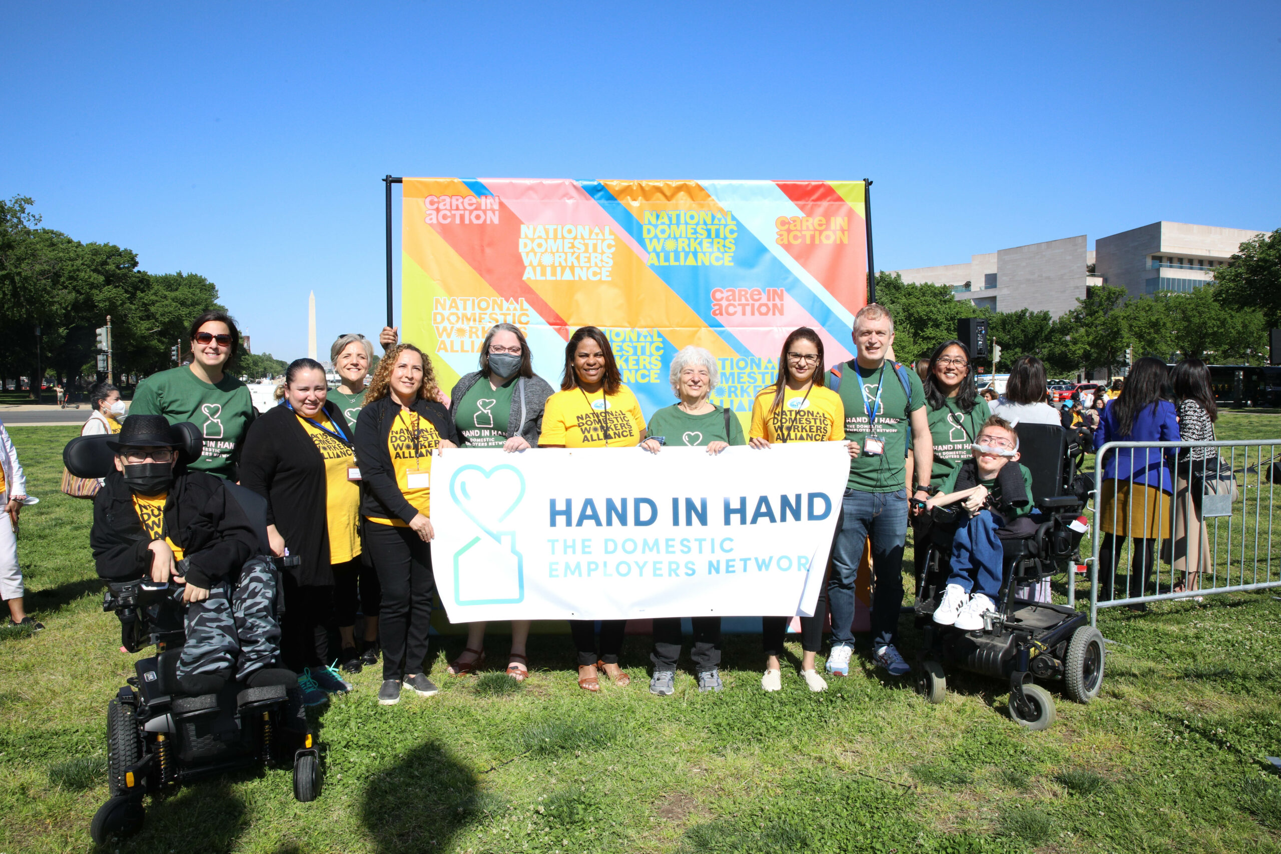 Group of people standing and sitting in wheelchairs while holding Hand in Hand banner, outside.