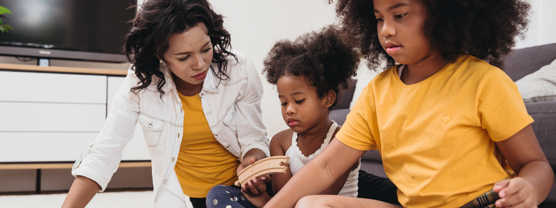 Two children sitting while concentrating on a task, along with their nanny who is helping them. 