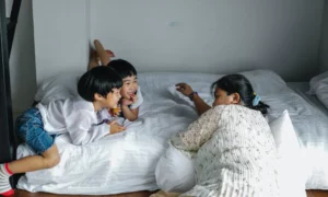 A women wearing a white shirt laying on bed is facing two smaller children while they all laugh together.