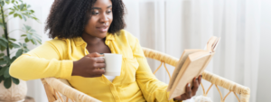 A black woman with a yellow shirt, holding a white mug while reading a book.