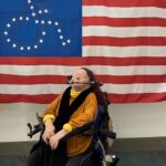  white woman wearing a headband, orange sweater over a yellow turtleneck, black scarf and glasses, sseated in a personalized wheelchair. Behind her is the flag of the United States, with stars in the shape of the wheelchair-accessible symbol. 