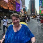 Janine, a white woman with short blonde hair sitting in a wheelchair in the middle of Times square. While wearing a blue watercolored print shirt, and a blue necklace. 