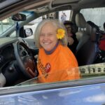 Julie Farrar, a white woman with silver hair and a sunflower behind her hair, wearing a bright orange shirt, sitting on the driver side of a car while smiling 