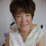 A headshot of a white woman with short brown hair sitting in a wheelchair, with a sparkly sequenced white dress with a white sash, smiling. 