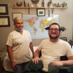 Middle-aged woman of color, standing next to Keith, a white man with short brown hair and beard, wearing a white t-shirt, sitting in a wheelchair smiling widely. 