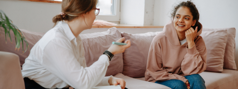White women in a white button up shirt and glasses holding a pen and speaking to another woman of color wearing a sweat shirt. They are both sitting on a couch.