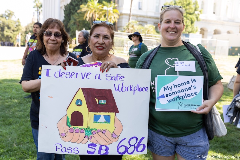 Two women of color hold signs that read "I deserve a safe workplace. Pass SB 686" They stand next to a white woman who holds a sign that reads "My home is someone's workplace"