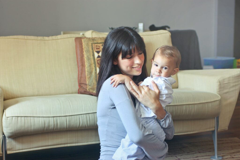 A nanny holds a toddler in her arms in front of a couch.