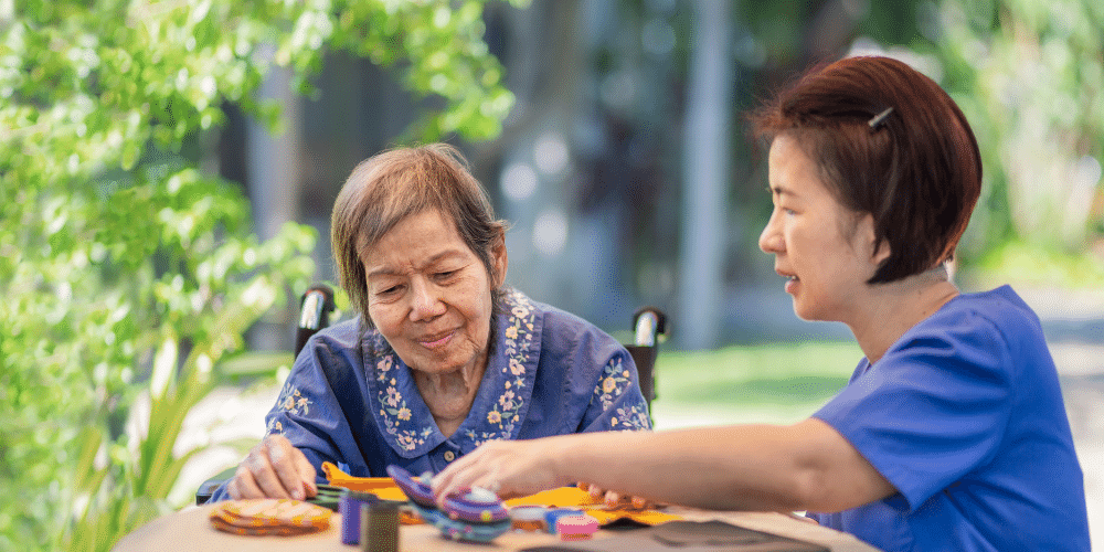 Asian woman wearing a blue nursing shirt sitting next to an older asian woman in a wheelchair. The two are playing a game.