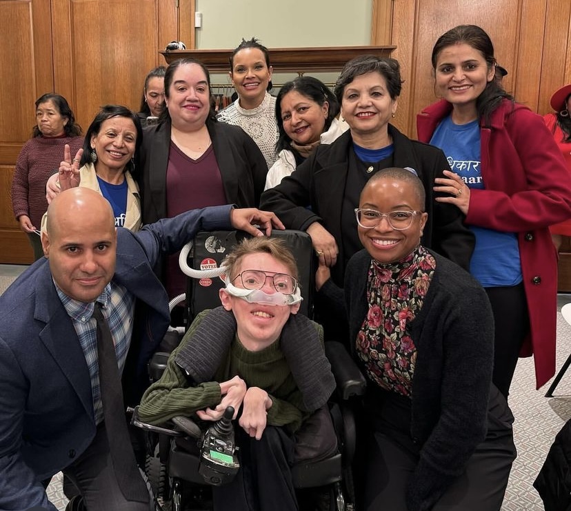 Steve Way, white man wearing glasses, seated in a wheelchair, surrounded by Jenn Stowe, Virgilio Aran and several National Domestic Workers Alliance and Adhikaar staff and members of various races, ethnicities and genders.