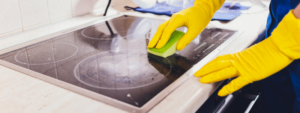 A hand wearing yellow rubber gloves wiping an electric stove with a green sponge.