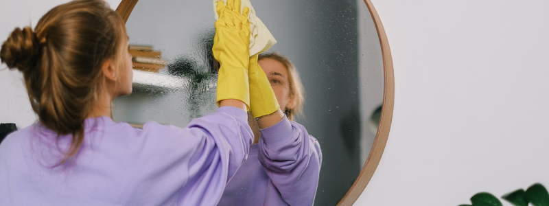 A white woman wearing yellow rubber gloves wiping a round glass mirror.