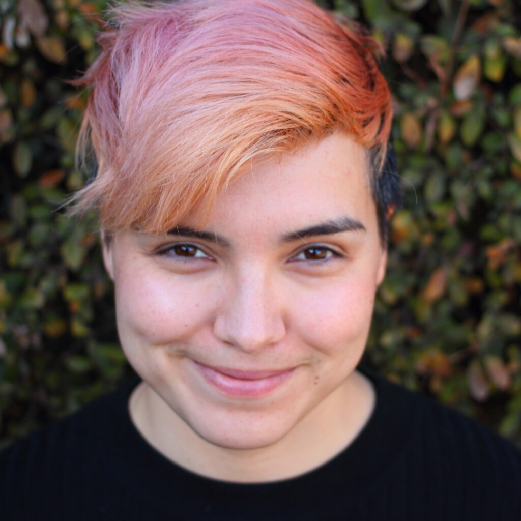 A headshot of a smiling non-binary Latine with short, pink hair wearing a black shirt in front of a green, leafy wall