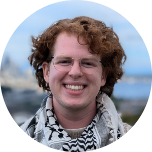 a white person with curly red hair, stands in front of a view of downtown Seattle. They are wearing a tan and black sweater and white and black kufiya.