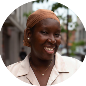 A headshot of a black woman wearing a head tie, that is tied into a back bun, smiling while on a sidewalk.