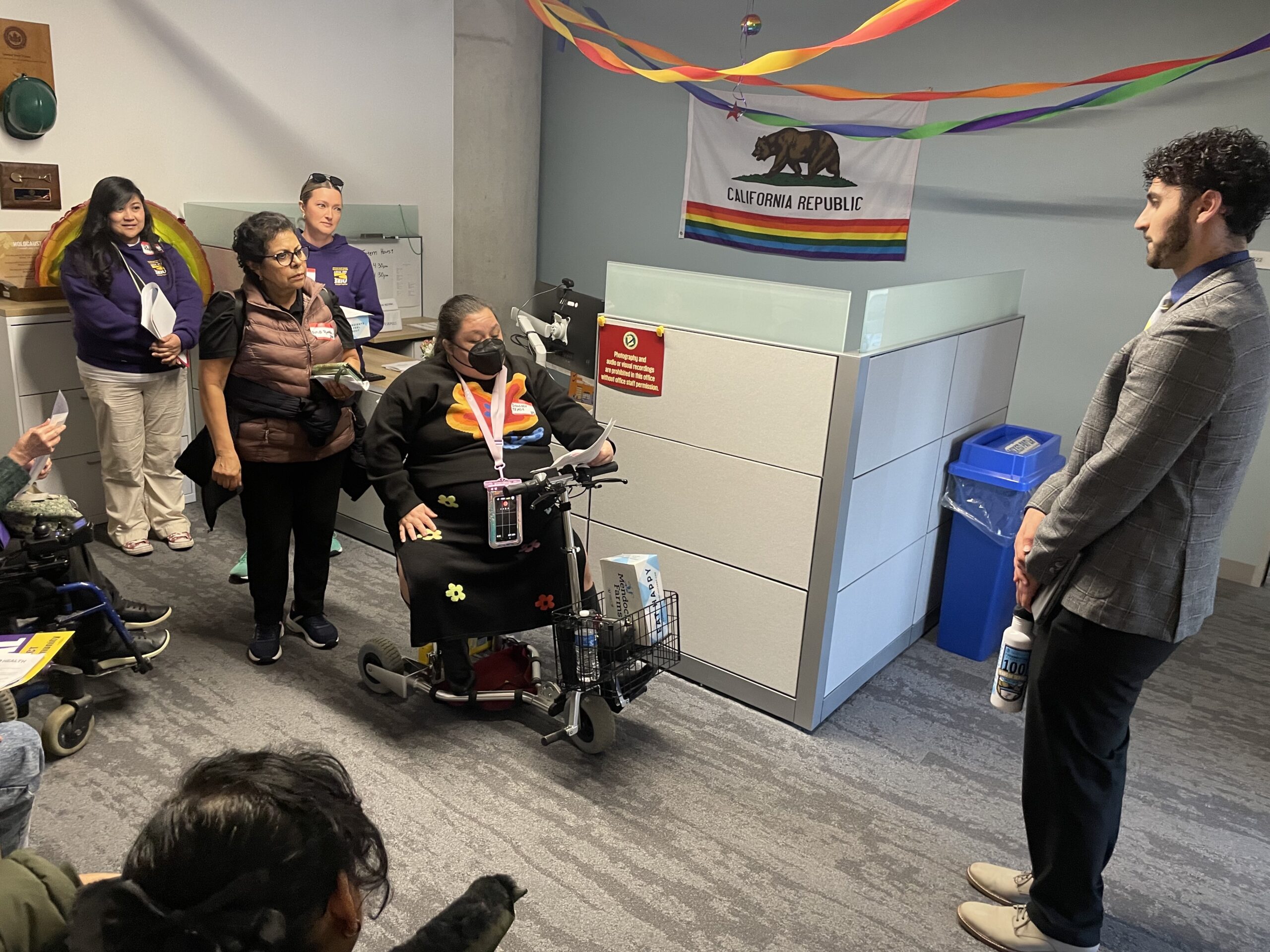 A person in a power wheelchair speaking to a legislative staffer in an office. Several advocates stand nearby, some using a walker and other mobility devices. A California flag with a rainbow stripe hangs on the wall.